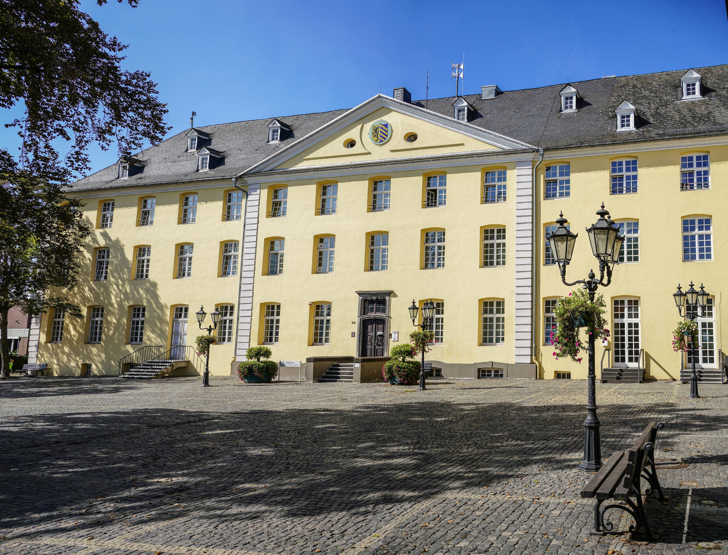 Gelbes historisches Gebäude Rathaus Brüggen mit Treppenaufgang zur braunen Eingangstür am gepflasterten Kreuzherrenplatz, Ausschnitt von Baumkrone, Schatten des Baumes auf Platz, auf dem Platz vereinzelt Straßenlaternen mit Hängepflanzen und Sitzbänke